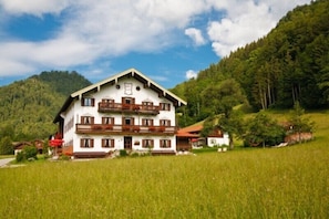 Un grand bâtiment à deux étages, doté d’un balcon à l’étage supérieur, entouré d’un paysage verdoyant et de collines.