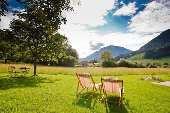 Un espace extérieur verdoyant avec deux chaises en bois et une table, un grand arbre et une montagne en arrière-plan.