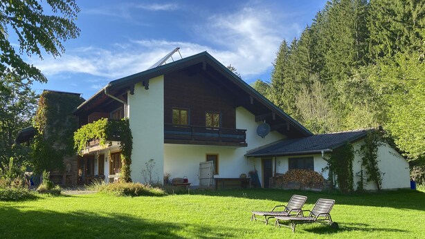 Eine zweistöckige Chalet mit Balkon, einem kleinen Anbau und einer Rasenfläche mit zwei Liegestühlen.