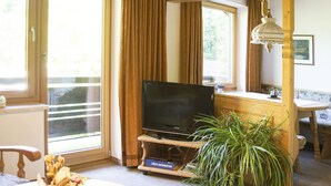 Ein Zimmer mit Fernseher auf einem Holzboden, einer Topfpflanze, einem Schreibtisch mit Lampe und einem fenster mit Vorhängen.