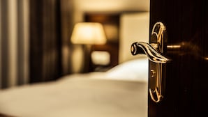 A hotel room with a wooden door, a bed, and a nightstand with a lamp.