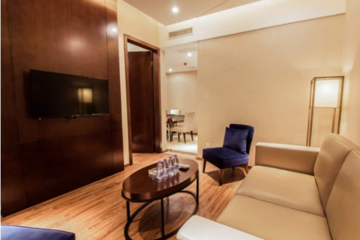 Una habitación de hotel moderna con una televisión de pantalla plana montada en la pared, una mesa de centro redonda de madera, un sofá color beige y un sillón azul.
