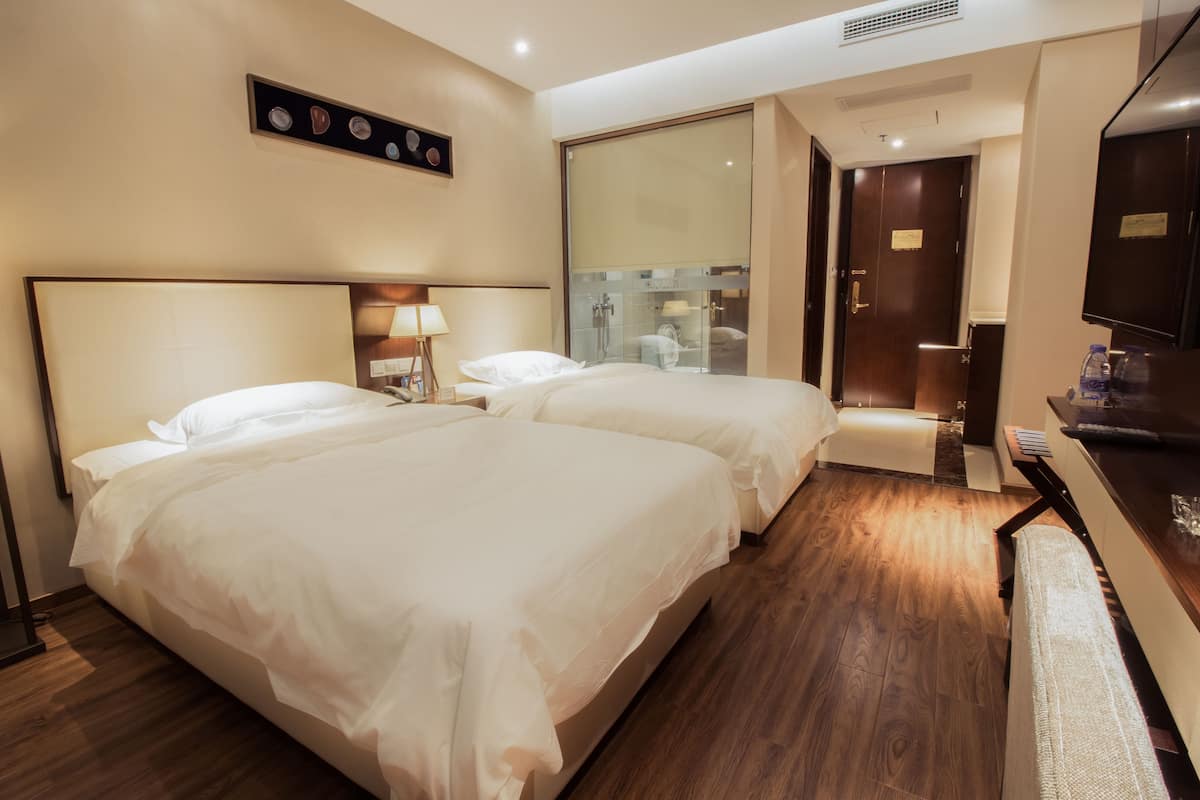 Habitación de hotel con dos camas, un escritorio, un televisor y un baño visible a través de una puerta de cristal.