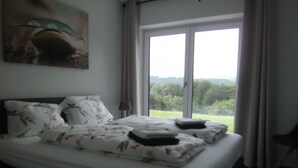 A neatly made bed with floral bedding, a painting on the wall, and a view of greenery through the window. - Moselle and Hunsrück - Enjoy the wine region and nature (Morbach)