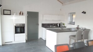 A modern kitchen with white cabinets, a built-in oven, and a large island with a sink. There are two chairs at the island and a dining table in the foreground. - Moselle and Hunsrück - Enjoy the wine region and nature (Morbach)