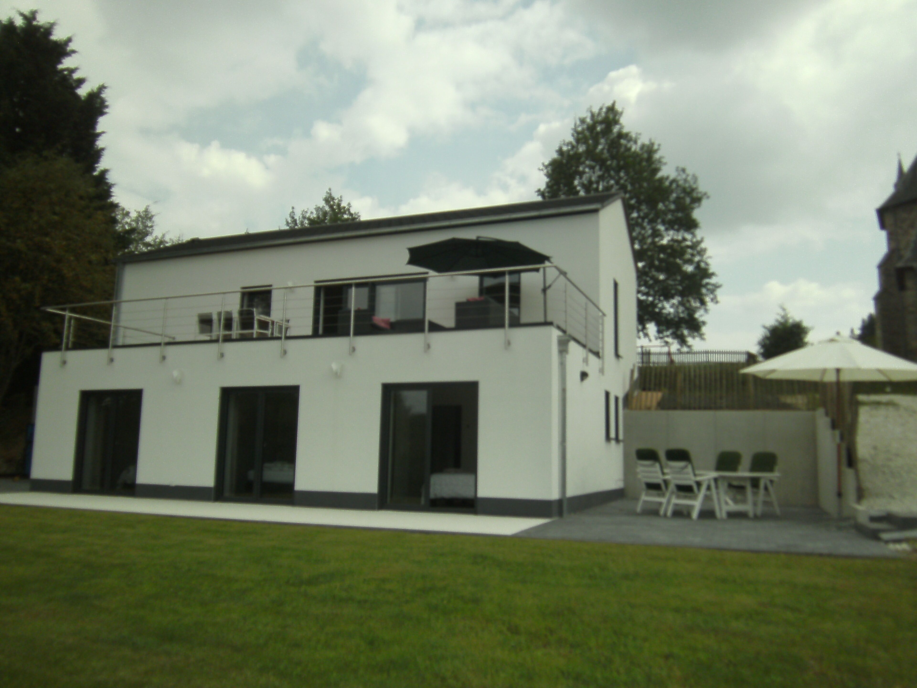 A modern two-story building with a flat roof, large windows, and a balcony. There is a white umbrella and outdoor seating area.