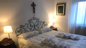 Een netjes opgemaakt bed met bloemenbeddels, twee bedlampjes, een kruis aan de muur en een ingelijste foto.