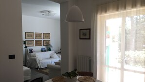 Een kamer met een eettafel, gedekt met geweven placemats, een plant in pot en een wit tafelkleed. Achtergrond toont een bed met een witte dekbedovertrek en kussens, een radiator en beelden in lijsten aan de muur.