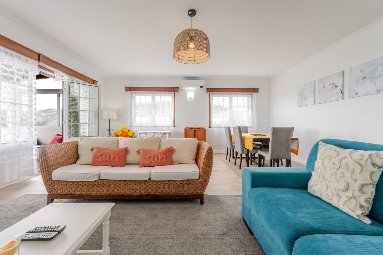 A living room with a wicker sofa, a blue sofa, a dining area, and a large window.