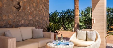 A wicker sofa and chair set with a stone wall backdrop, a round table with drinks, and a view of greenery and palm trees.