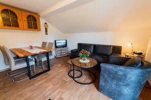 Ein Wohnzimmer mit einem Holzesstisch, einer schwarzen Couch, einem kleinen runden Couchtisch mit Blumenvase und einem Fernseher auf einem Lowboard.