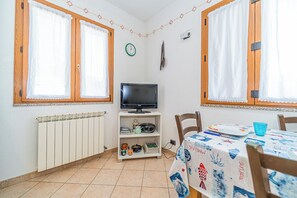 Ein kleiner, gut beleuchteter Raum mit zwei Fenstern, einem Heizkörper, einem Fernseher auf einem Regal und einem Tisch mit Tischdecke.