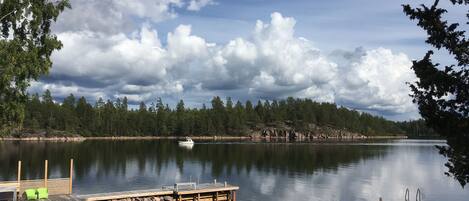 Un lac paisible, avec un quai et un bateau, entouré d’arbres et surplombé d’un ciel nuageux.