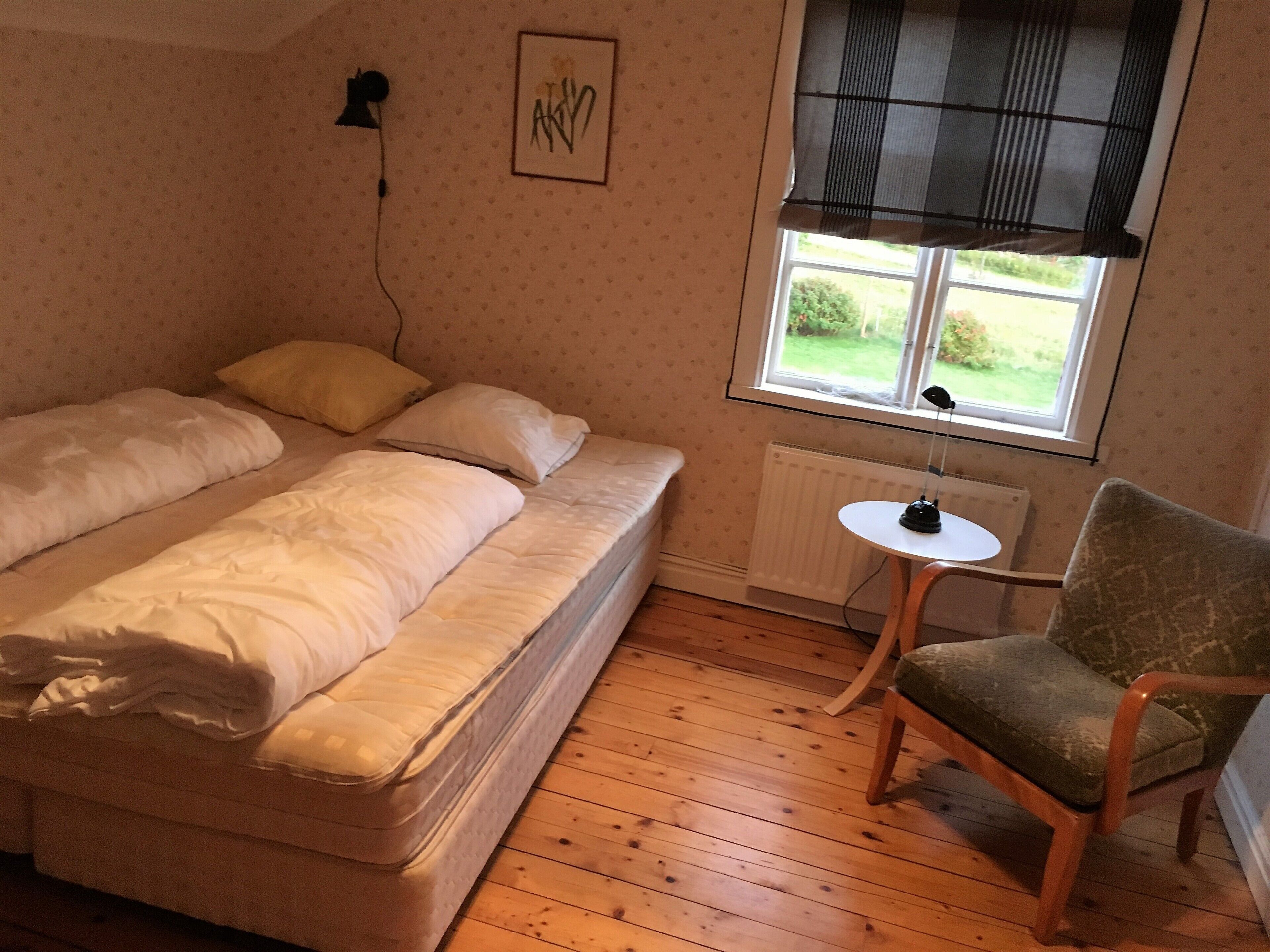 A small, cozy room with a bed, a chair, a small table, and a window with a view of greenery outside.