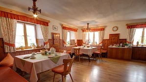 Un comedor con mesas y sillas de madera, una cocina con armarios de madera, y ventanas grandes con cortinas.