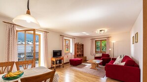Ein geräumiges Wohnzimmer mit Holzboden, einem roten Sofa, einem Esstisch mit Stühlen und einem Fernseher auf einem Holzschrank.