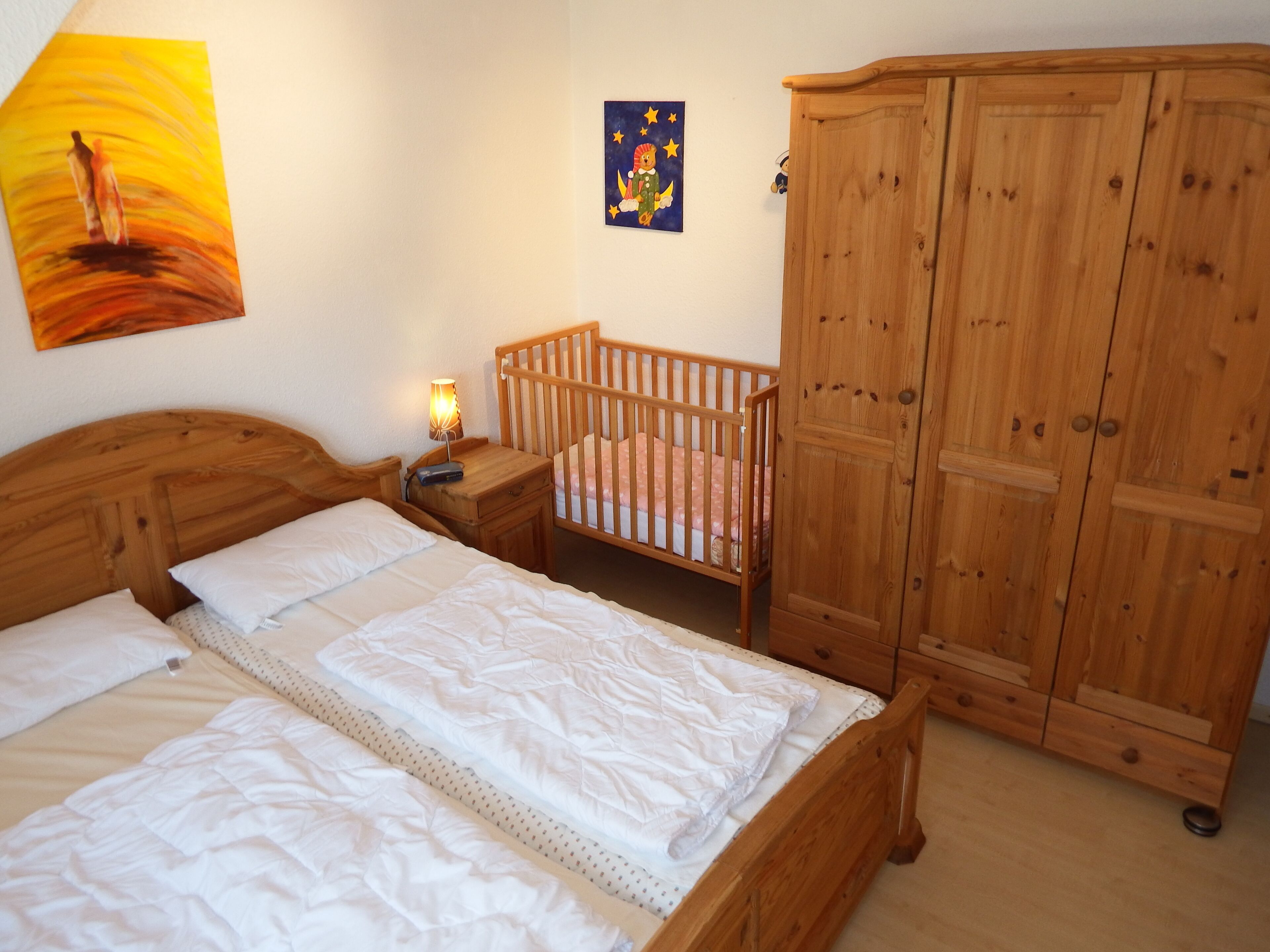 A bedroom with a wooden bed, a crib, a wardrobe, and a nightstand with a lamp.
