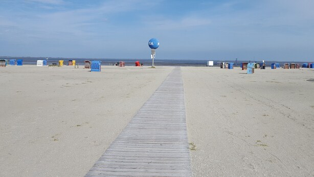 Una playa con un paseo marítimo de madera que conduce al mar, con varias cabañas de playa azules y rojas.