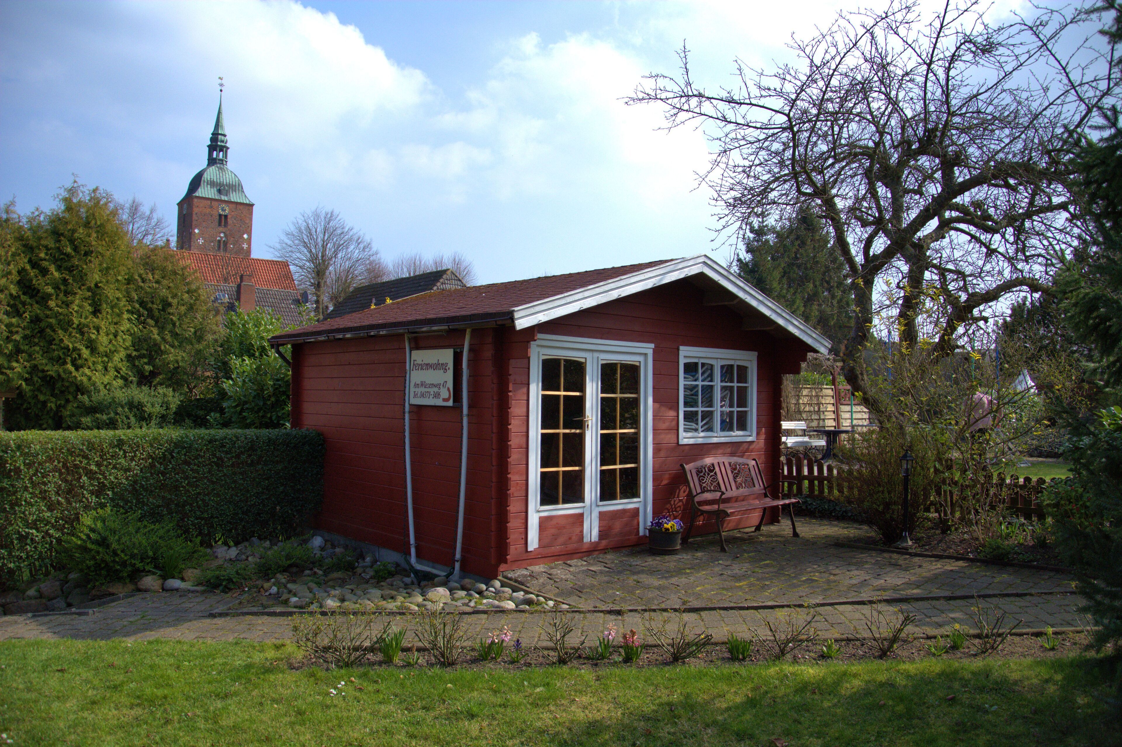 A small red wooden building with a white door and windows, surrounded by a garden with a bench and a tree.