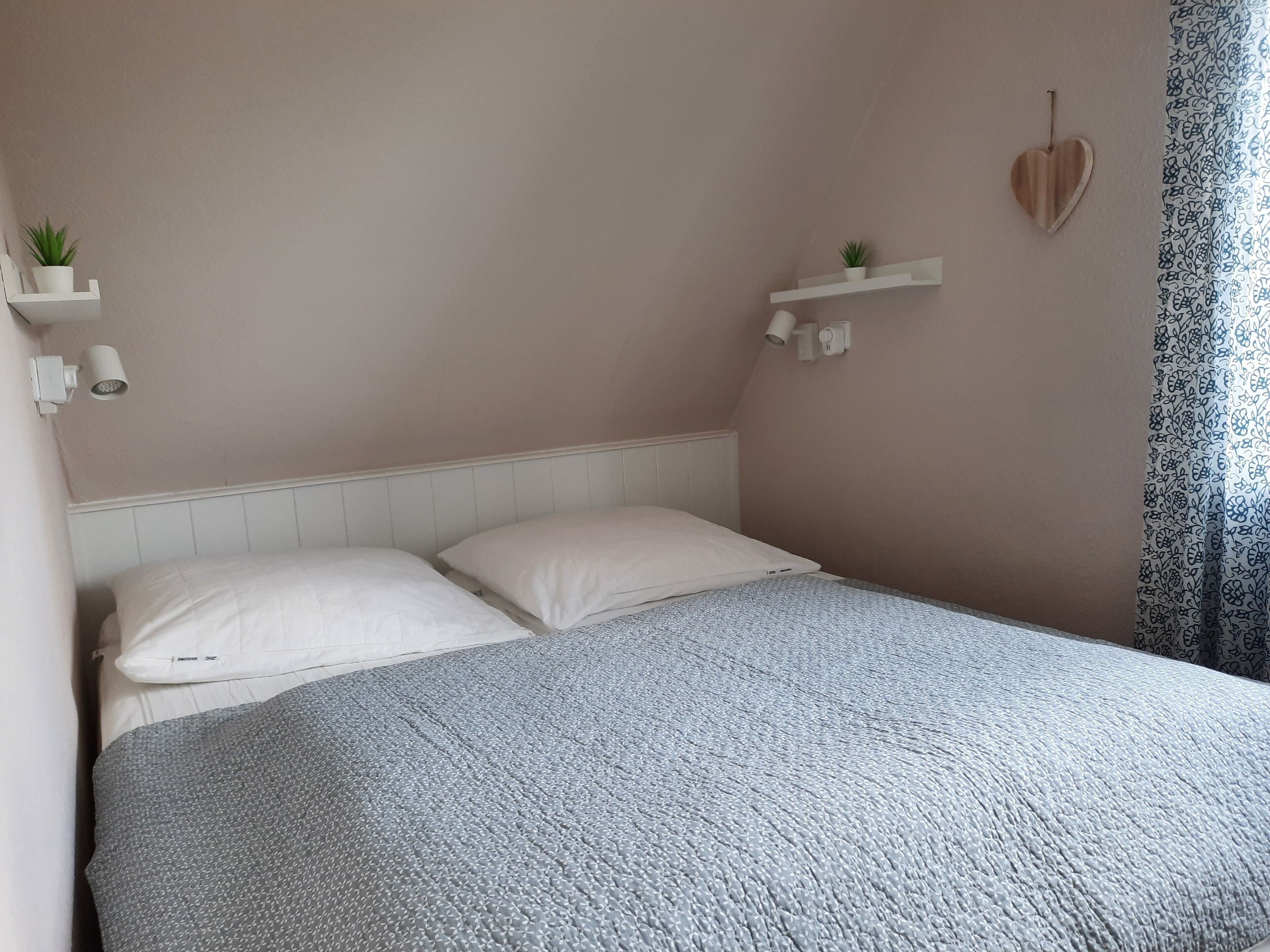 A bedroom with a bed, two white pillows, a grey textured blanket, and two small potted plants on white floating shelves.