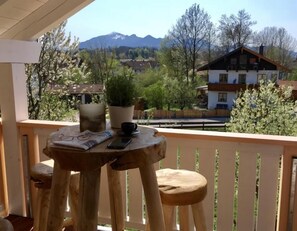A wooden table with a potted plant, a cup, and a smartphone on a balcony with a view of houses and mountains.