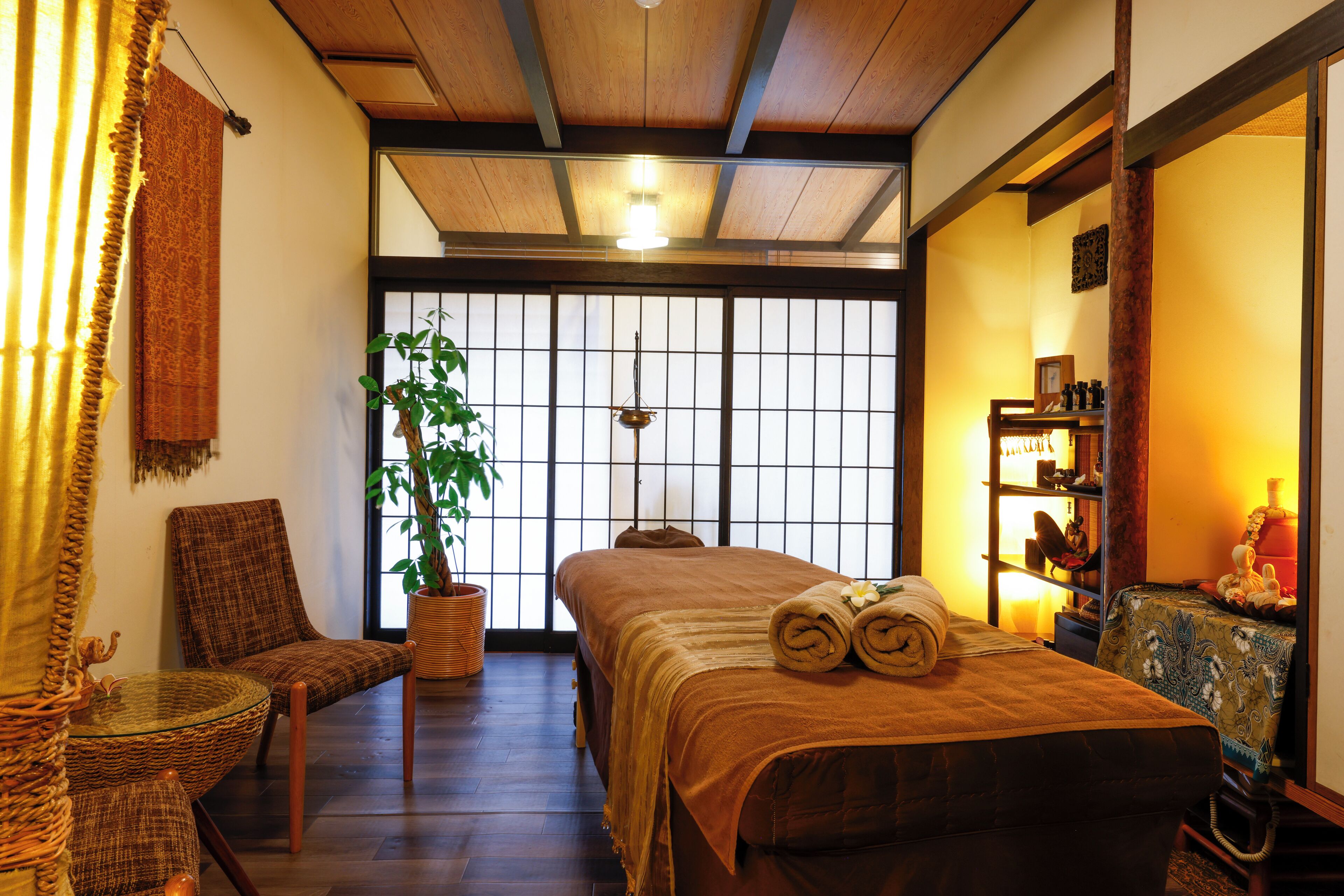 Una habitación tradicional japonesa con suelo de tatami, una cama baja con manta marrón, una silla de madera, una planta en maceta y una puerta corredera con diseño de cuadrícula.