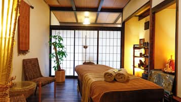Una habitación tradicional japonesa con suelo de tatami, una cama baja con manta marrón, una silla de madera, una planta en maceta y una puerta corredera con diseño de cuadrícula.