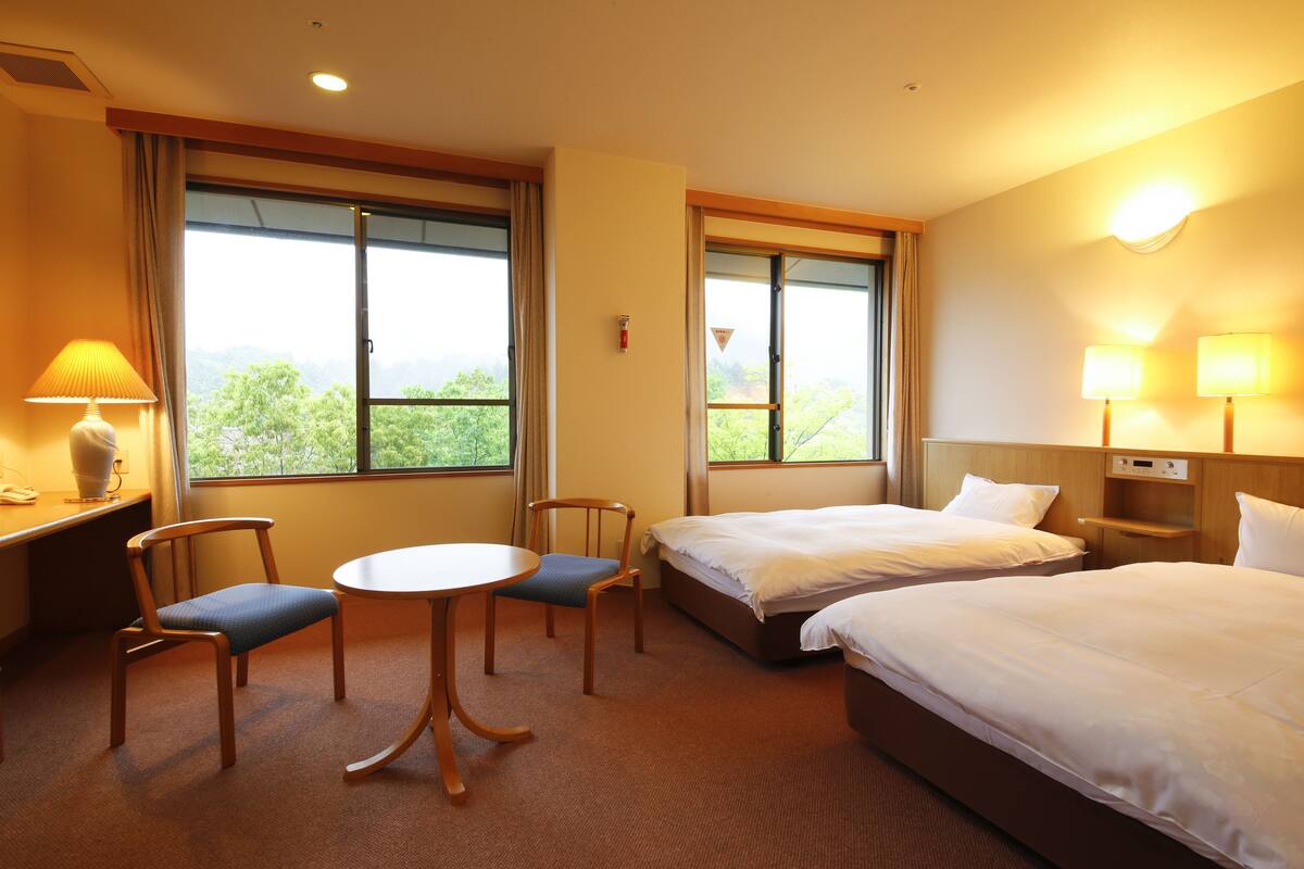 A hotel room with two single beds, a small table with chairs, a desk with a lamp, and large windows offering a view of greenery.