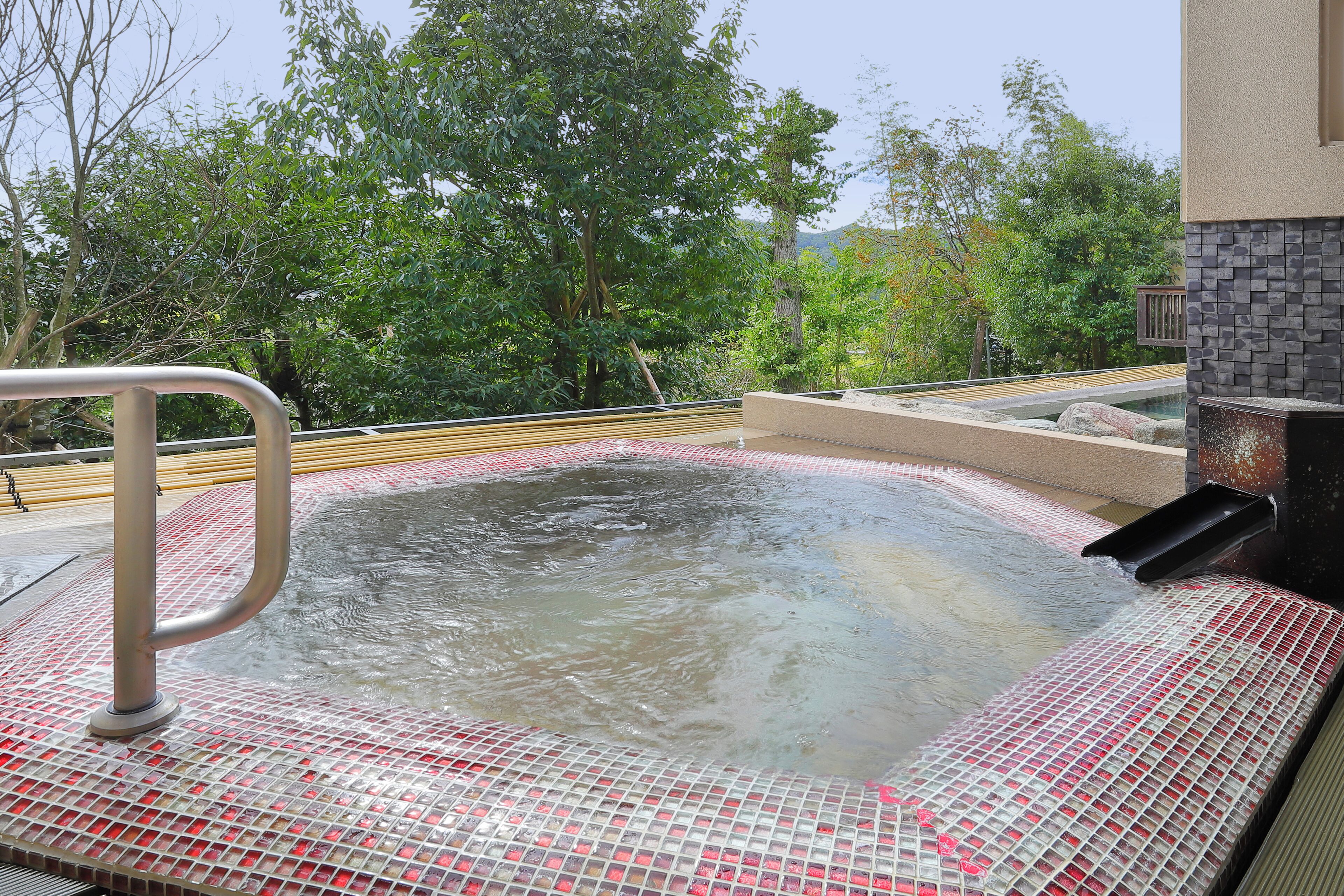 Un jacuzzi con borde de azulejos y barandilla de metal, rodeado de follaje.