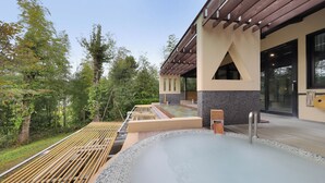 Un espace avec un bain à remous, une terrasse en bois, un coin salon abrité et un bâtiment doté de grandes fenêtres et d’une façade en pierre.