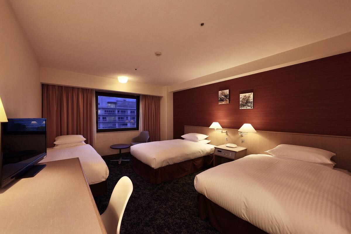 A hotel room with two single beds, a desk with a computer, a TV, and a window with curtains.