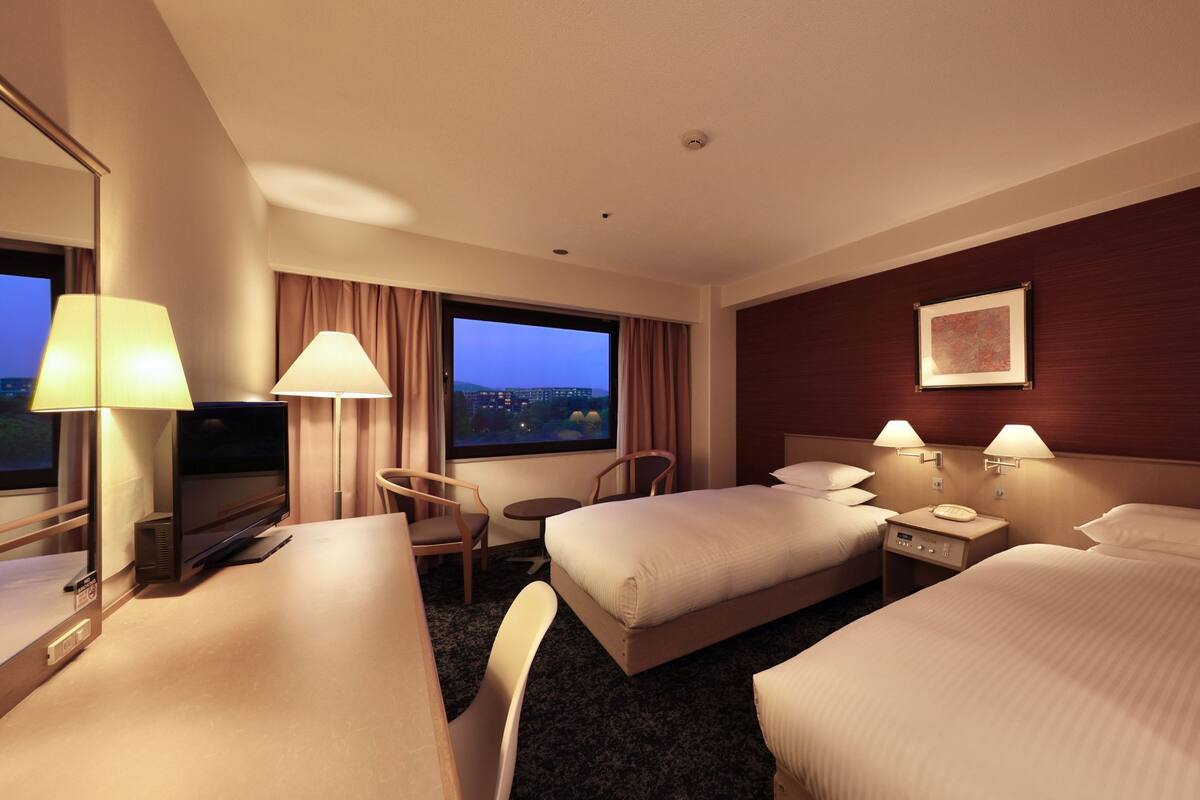 A hotel room with two beds, a desk, a chair, a small table, and a window with a view of the cityscape.