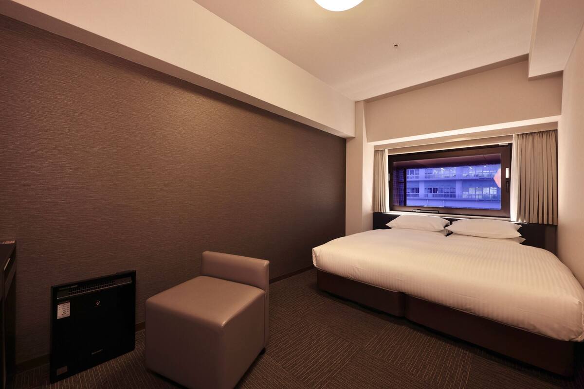 A hotel room with a bed, a television, a small seating area, and a window with a view of buildings.