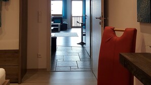 Una habitación con piso de madera, una silla roja, un banco de madera, un cuadro en la pared y vista a un comedor a través de una puerta abierta.