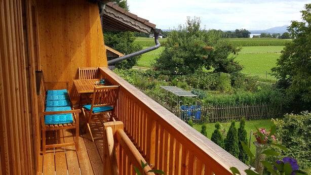 Una terraza de madera con mesa y sillas, un canalón metálico y vistas a un jardín exuberante y colinas lejanas.