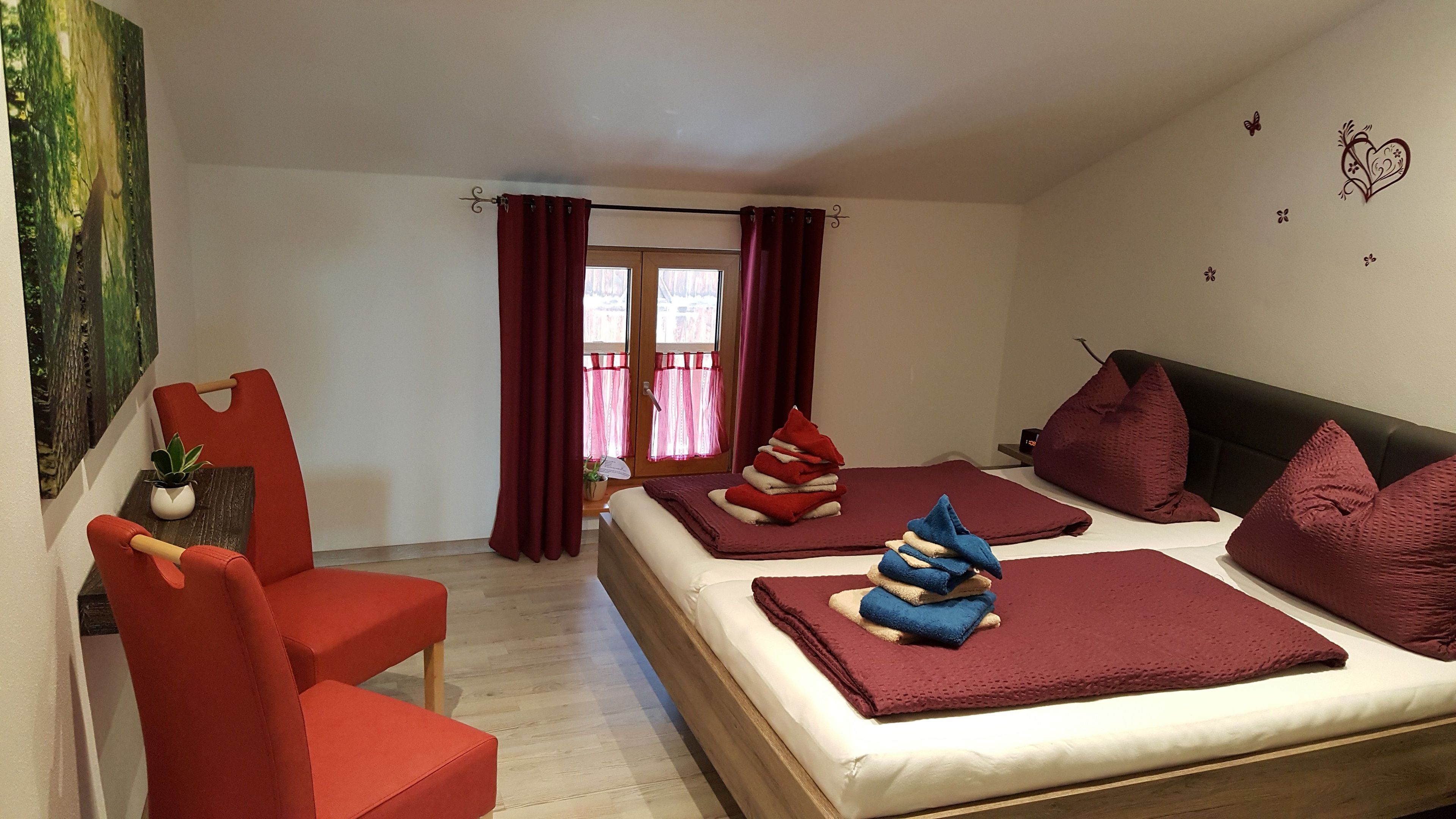 Habitación de hotel con una cama, dos sillas rojas, una mesita con una planta y un cuadro en la pared.