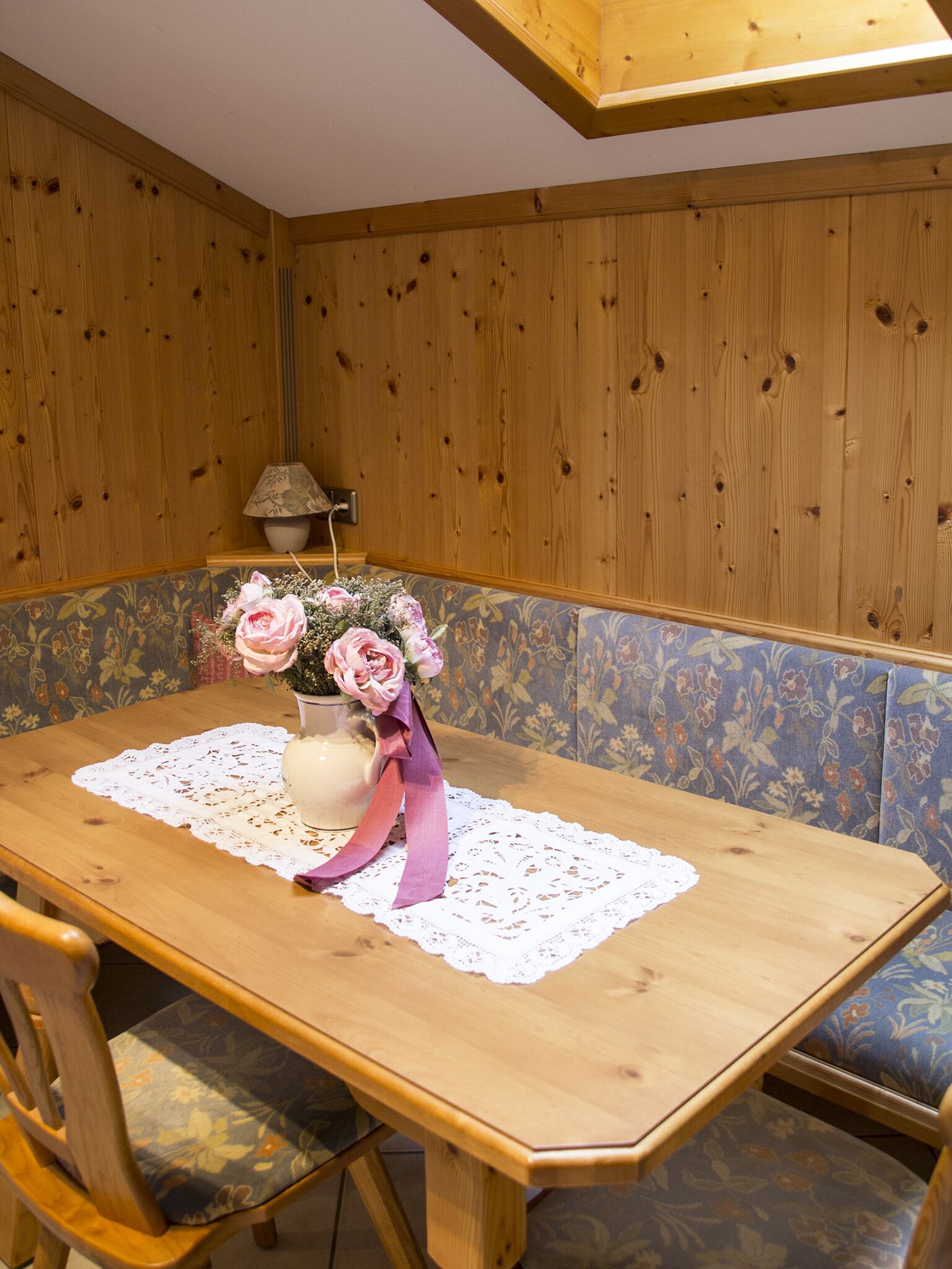 A wooden dining table with a lace tablecloth, a vase with pink flowers, and a bench with floral upholstery.
