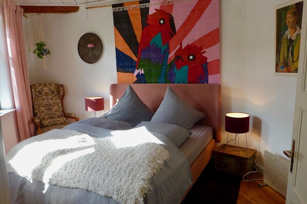 Une chambre à coucher avec un lit, une chaise, une plante suspendue, une tapisserie colorée et un tableau au mur.