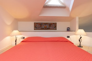 A bedroom with a skylight, a bed with a red bedspread, two bedside lamps, and a decorative wall piece.