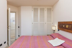 Un dormitorio con cama, cabecera de madera, mesita de noche con lámpara y puerta que da a otra habitación.