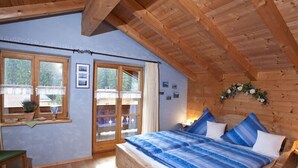 Une chambre avec un plafond en bois, un grand lit avec une literie bleue, une fenêtre donnant sur un beau paysage et une petite table sur laquelle est posée une plante.