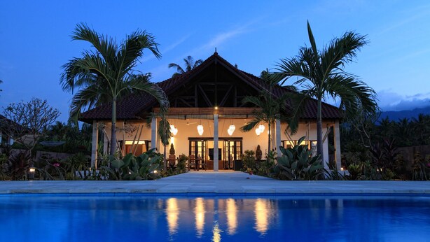 Uma casa de dois andares com piscina na frente, cercada por palmeiras e plantas tropicais.