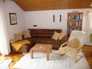 Ein gemütliches Wohnzimmer mit einem braunen Sofa, einem Holztisch, einem mit Fell bezogenen Sessel, einem Bücherregal voller Bücher und einem weißen, flauschigen Teppich.