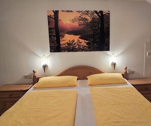 A bed with two yellow pillows, a wooden headboard, and a wall-mounted painting of a sunset over a lake.