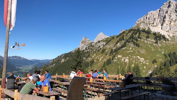 Vista de la montaña con mesas y bancos de picnic de madera, una bandera y un cielo azul despejado.