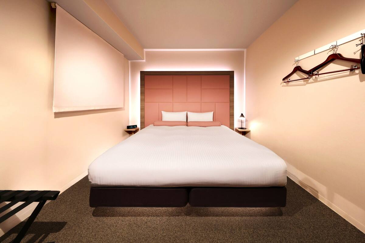 A modern hotel room with a large bed, a bedside table with a lamp, a wall-mounted coat rack, and a large window.