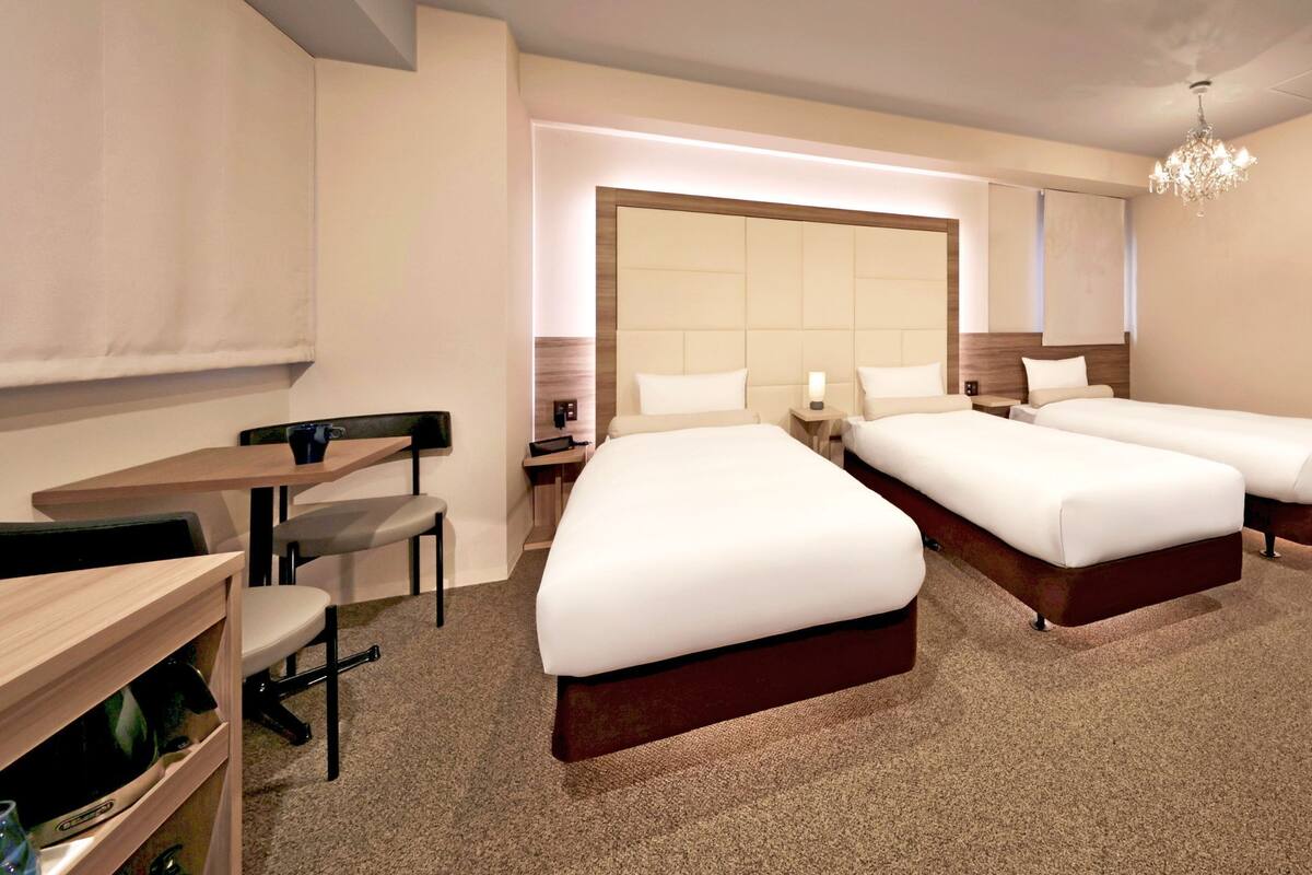 A hotel room with two beds, a desk, a chair, a small table, a chair, a bedside table, a lamp, a heater, and a chandelier.