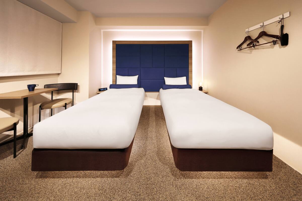 A hotel room with two single beds, a desk, a chair, a clock, and a coat rack.