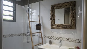 Une salle de bain avec une douche, un lavabo, un miroir avec un cadre en bois et une petite fenêtre.