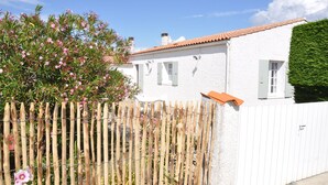 Un bâtiment blanc avec un toit recouvert de tuiles rouges, une clôture en bois et un jardin bien entretenu où poussent des fleurs roses.